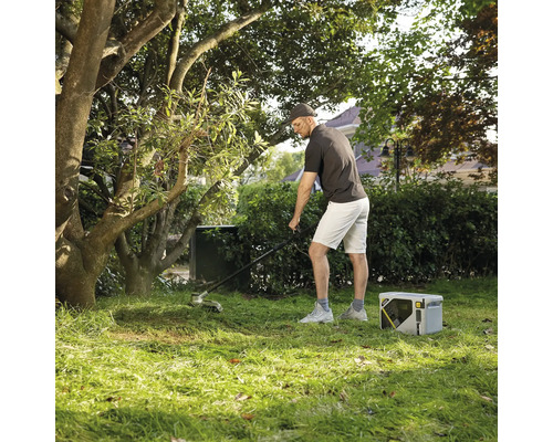 Homme effectuant l'entretien du jardin avec un coupe-bordure électrique et une station électrique portable sur une pelouse sous des arbres.