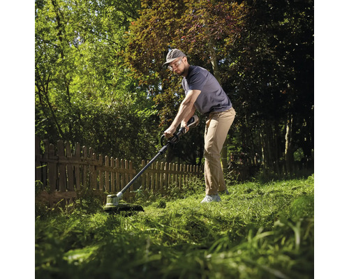 Un homme utilise un coupe-bordures près d'une clôture en bois dans un jardin vert et ensoleillé.