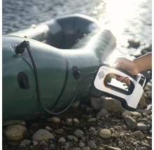 Une main tient une pompe à air électrique portable avec Lithell Logo gonflant un bateau pneumatique sur une rive rocailleuse au bord de l'eau.