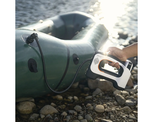 Une main tient une pompe à air électrique portable avec Lithell Logo gonflant un bateau pneumatique sur une rive rocailleuse au bord de l'eau.