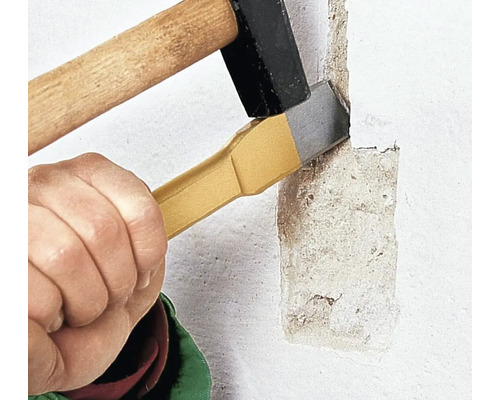 Une main maintient un burin plat contre un mur crépi pendant qu'un marteau frappe l'extrémité du burin pour retirer l'enduit.
