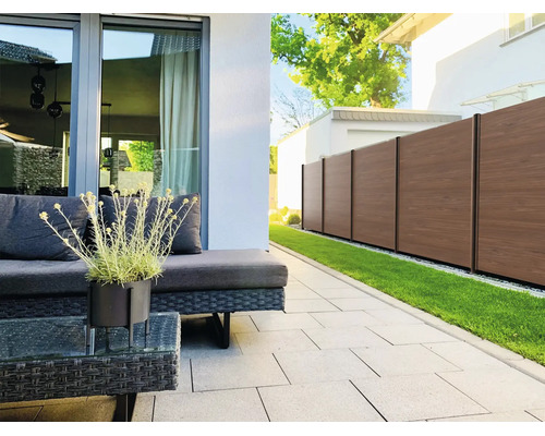 Terrasse moderne avec meubles de jardin en rotin foncé, pavés gris clair et clôture de protection brune à côté d'une pelouse.