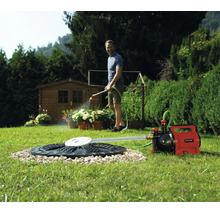Un homme arrose des plantes avec une pompe de jardin Einhell raccordée à une citerne dans la pelouse. Un abri de jardin figure en arrière-plan.