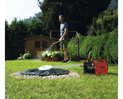 Un homme arrose des plantes avec une pompe de jardin Einhell raccordée à une citerne dans la pelouse. Un abri de jardin figure en arrière-plan.
