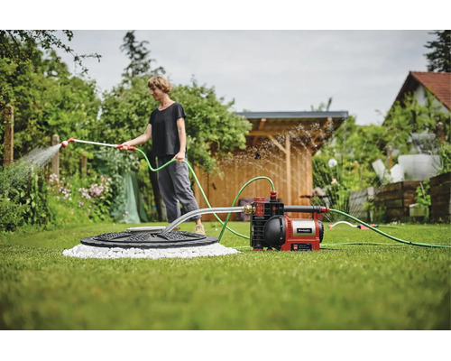 Einhell Gartenpumpe auf Rasen, verbunden mit Wasserquelle und Schlauch zur Bewässerung von Pflanzen in einem hellen, organisierten Garten.