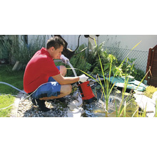 Un homme installe une pompe à eau chargée Einhell rouge avec raccord de tuyau dans un bassin de jardin planté.