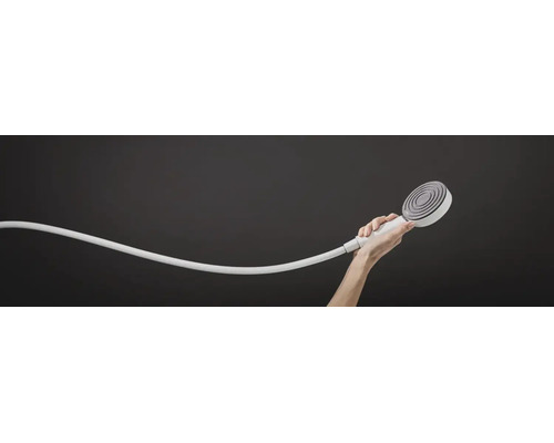 Une main tient une douchette ronde avec flexible de douche devant un fond neutre