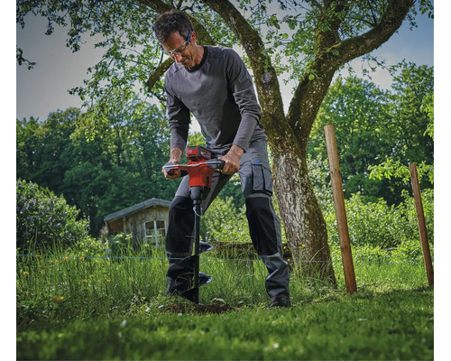 Ein Handwerker nutzt einen akkubetriebenen Erdbohrer im Garten für Erdarbeiten in der Wiese.
