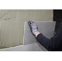 Artisan avec des gants de travail pose un carreau sur un mur enduit de colle à carrelage