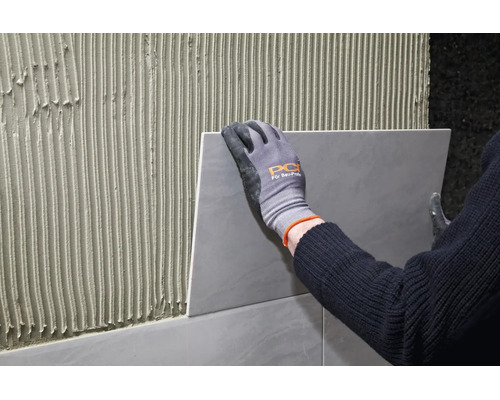 Artisan avec des gants de travail pose un carreau sur un mur enduit de colle à carrelage