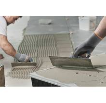 Artisan appliquant de la colle à carrelage avec une truelle dentée sur un sol pour la pose de carrelage.