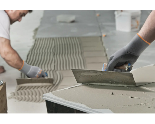 Artisan appliquant de la colle à carrelage avec une truelle dentée sur un sol pour la pose de carrelage.