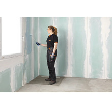 Une femme en tenue de travail peint un mur en plaques de plâtre traité avec un apprêt dans une pièce à rénover à l'aide d'un rouleau.