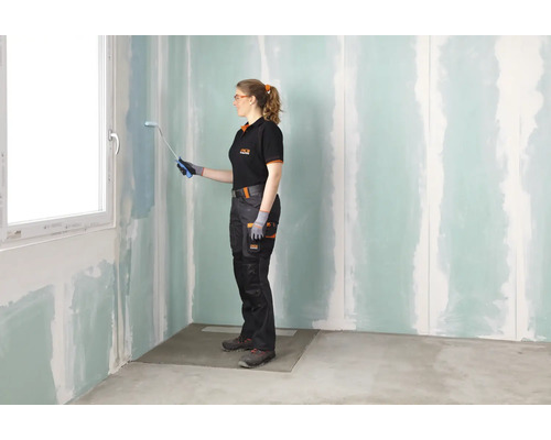 Une femme en tenue de travail peint un mur en plaques de plâtre traité avec un apprêt dans une pièce à rénover à l'aide d'un rouleau.