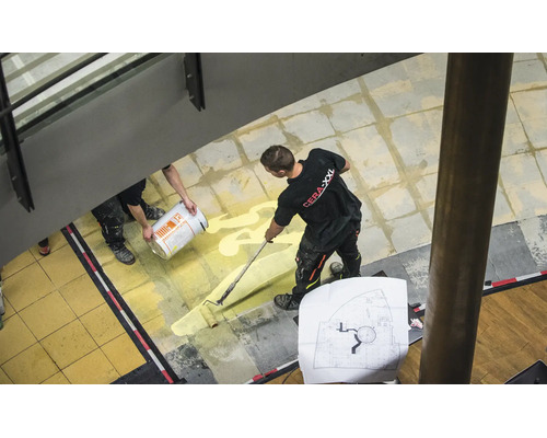 Artisan appliquant une couche de fond avec un rouleau à peinture sur un sol carrelé en intérieur.