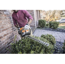 Eine Person schneidet eine Hecke mit einer motorbetriebenen Stihl Heckenschere an einer Backsteinmauer in einem Garten.