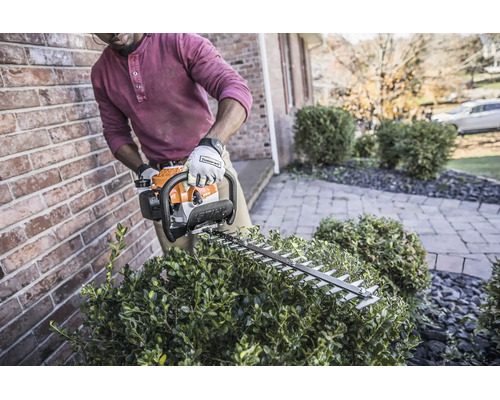 Eine Person schneidet eine Hecke mit einer motorbetriebenen Stihl Heckenschere an einer Backsteinmauer in einem Garten.