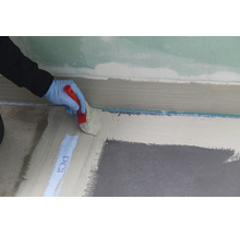 Une main gantée applique au pinceau une étanchéité liquide sur le sol le long d'un mur.
