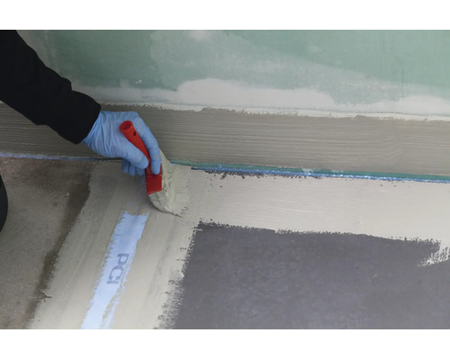 Une main gantée applique au pinceau une étanchéité liquide sur le sol le long d'un mur.