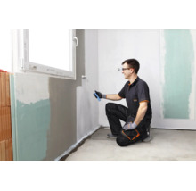 Un artisan peint un mur intérieur sous une fenêtre avec un rouleau de peinture.