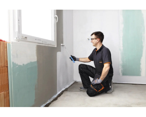 Un artisan peint un mur intérieur sous une fenêtre avec un rouleau de peinture.