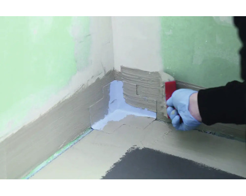 Main avec gant appliquant une bande d etancheite dans un angle de mur pour une etancheite sous carrelage.