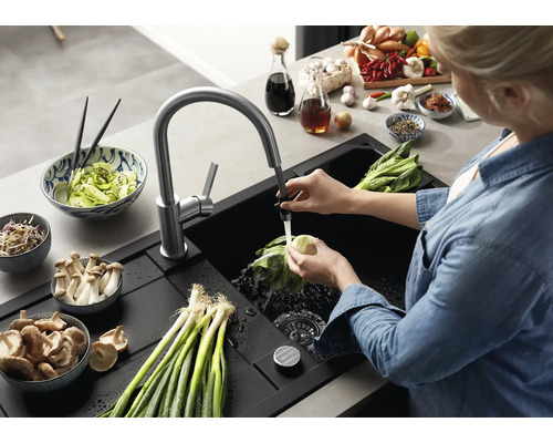 Une personne lave des légumes dans un évier encastrable noir moderne avec mitigeur de cuisine extractible dans une cuisine organisée.