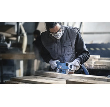 Artisan ponçant une planche en bois avec une ponceuse à bande Bosch dans un atelier, portant des vêtements de protection et un masque.