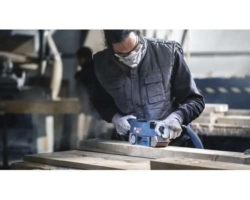 Artisan ponçant une planche en bois avec une ponceuse à bande Bosch dans un atelier, portant des vêtements de protection et un masque.