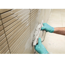 Handwerker mit Handschuhen verfugt eine geflieste Wand mit einer Fugenkelle.