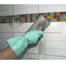 Une main avec un gant de protection étale du mortier de jointoiement avec une truelle sur un mur de carrelage avec une bordure en mosaïque.