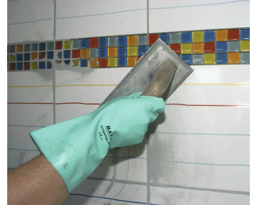 Une main avec un gant de protection étale du mortier de jointoiement avec une truelle sur un mur de carrelage avec une bordure en mosaïque.