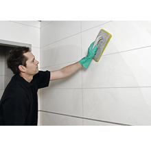 Artisan réalisant le jointoiement de carreaux muraux avec une taloche à joint et des gants de protection dans une pièce carrelée.