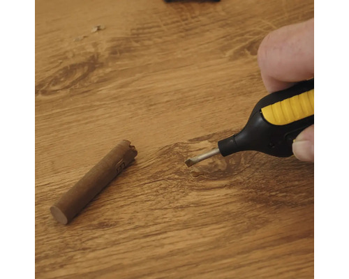 Hand der einen Wachsschmelzer über einer Holzoberfläche hält neben einem Wachsstab zur Reparatur von Bodenbelägen