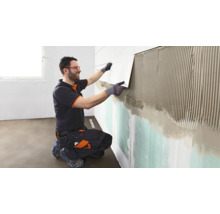 Un artisan portant des vêtements de travail et des lunettes de protection pose un carreau sur le mur enduit de colle à carrelage.