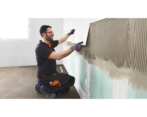 Un artisan portant des vêtements de travail et des lunettes de protection pose un carreau sur le mur enduit de colle à carrelage.