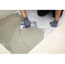 Artisan appliquant de la colle à carrelage au sol avec une spatule dentée pour la pose de carrelage.