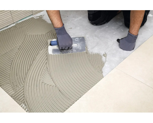 Artisan appliquant de la colle à carrelage au sol avec une spatule dentée pour la pose de carrelage.