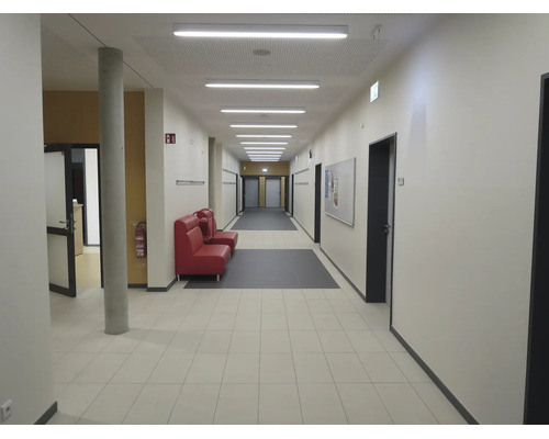 Couloir lumineux dans un bâtiment public avec sol carrelé, une colonne en béton, des sièges, des portes et un éclairage de plafond.