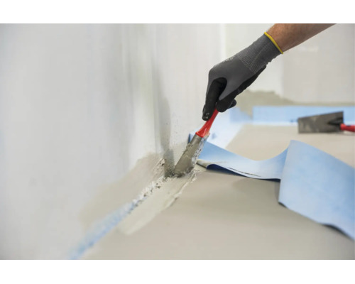 Main d un ouvrier portant des gants de protection appliquant du mortier hydrofuge au pinceau sur une jonction mur sol avec une bande d étanchéité