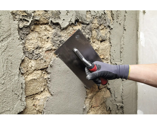 Une main gantée lisse du plâtre frais avec une truelle sur une surface de maçonnerie inégale.