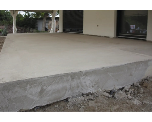 Une terrasse en béton fraîchement coulée devant une maison, la surface est lissée et les bords ne sont pas encore totalement durcis.