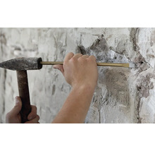 Artisan travaillant avec un marteau de mécanicien et un burin pour enlever de la maçonnerie sur un mur crépi.