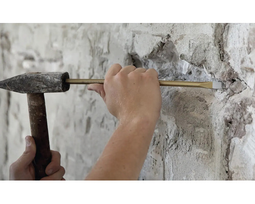 Artisan travaillant avec un marteau de mécanicien et un burin pour enlever de la maçonnerie sur un mur crépi.