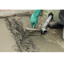 Un artisan lisse une chape de béton fraîche avec une brosse sur un sol à côté d'un tuyau d'évacuation.