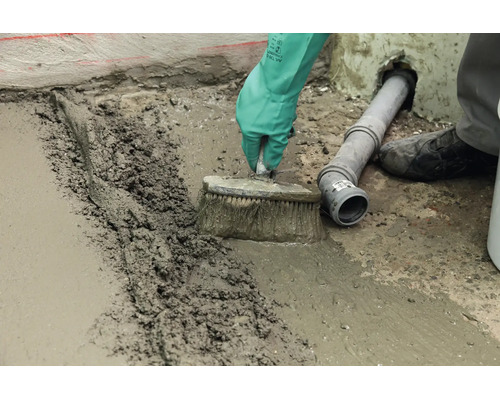 Un artisan lisse une chape de béton fraîche avec une brosse sur un sol à côté d'un tuyau d'évacuation.