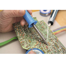 Une main soude un composant électronique sur un circuit imprimé vert à l'aide d'un fer à souder.