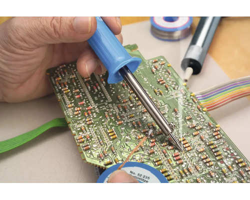 Une main soude un composant électronique sur un circuit imprimé vert à l'aide d'un fer à souder.