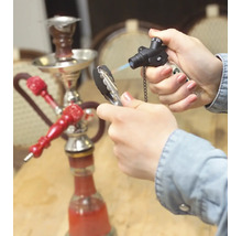 Eine Hand entzündet Shishakohle mit einem Gasbrenner, im Hintergrund steht eine Shisha auf einem Tisch.