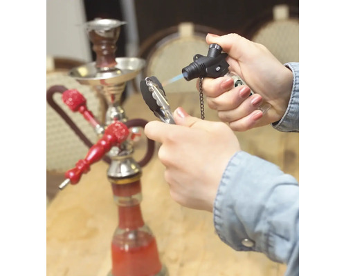 Eine Hand entzündet Shishakohle mit einem Gasbrenner, im Hintergrund steht eine Shisha auf einem Tisch.